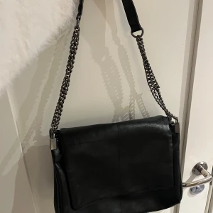 Svart axelväska Zara - Snygg svart axelväska i skinn med kedjedetaljer på axelremmen. Väskan har flera fack med dragkedja för praktisk förvaring. Perfekt för en stilren look.