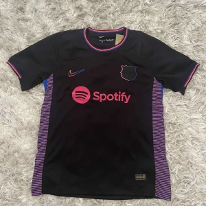 Svart fotbollströja från Nike - Snygg svart fotbollströja från Nike med korta ärmar. Tröjan har rosa och blå detaljer samt ett tryck av Spotify-loggan på framsidan. Perfekt för fotbollsfans som vill ha en stilren look.