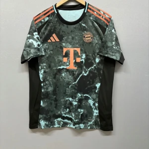 Svart och grön FC Bayern München fotbollströja - Snygg FC Bayern München fotbollströja från Adidas i svart och grön med orange detaljer. Tröjan har korta ärmar och ett unikt mönster över hela tyget. Perfekt för fans av laget!