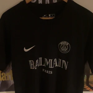 Svart t-shirt från Nike x Balmain - Snygg svart t-shirt från Nike i samarbete med Balmain. Den har en stilren design med Balmain Paris-logga och Nike swoosh på bröstet. Perfekt för dig som vill ha en exklusiv look med sportig touch.