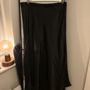 Svart långkjol från Zara - Elegant svart långkjol från Zara i ett glansigt material. Kjolen har en stilren design med knytband i midjan för justerbar.