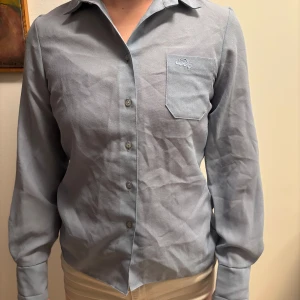 Ljusblå vintage skjorta med broderad detalj - Elegant ljusblå skjorta med långärmad design och knappar framtill. Skjortan har en broderad detalj på bröstfickan som ger en stilren touch. Perfekt för en klassisk look. Vintage 