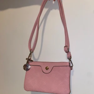 Rosa axelväska - Snygg rosa axelväska med präglat mönster och guldfärgade detaljer från Ulrika. Väskan har en justerbar axelrem och stängs med en tryckknapp. Insidan är fodrad med ett blommigt mönster och har en dragkedjeficka för extra förvaring.