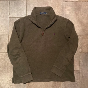Ralph Lauren Half Zip Medium - Säljer en stilren olivgrön Half/Quarter Zip från Polo Ralph Lauren med dragkedja och broderad logga i lila på bröstet. Perfekt för en avslappnad och snygg look. Storlek Medium i väldigt gott skick.