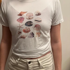 Vit t-shirt med snäckmotiv - Säljer en vit t-shirt med ett färgglatt snäckmotiv på framsidan. T-shirten är kortärmad och har en croppad passform, mjukt material 
