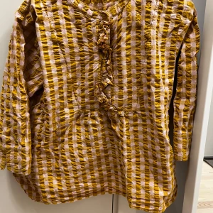 Gul och rosa rutig blus från Juna - Säljer en unik gul och rosa rutig blus från Juna. Blusen har trekvartsärmar och en dekorativ knytning framtill. Perfekt för att liva upp garderoben med lite färg och mönster!