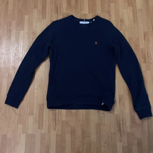 Mörkblå tröja med broderad detalj - Snygg mörkblå tröja med en liten broderad detalj på bröstet. Tröjan har långa ärmar och en klassisk rund halsringning. Perfekt för en stilren och avslappnad look. Stor i storleken 