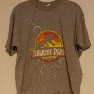 Grå Jurassic Park barn t-shirt - Cool grå t-shirt med Jurassic Park-logga i rött, gult och orange. Tillverkad i en bomull- och polyesterblandning. Perfekt för filmälskare! Storlek 122-128 