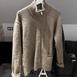 Beige kofta från Weekday - Säljer en stilren beige kofta från Weekday med dragkedja och hög krage. Koftan har långa ärmar och en praktisk ficka framtill. Perfekt för kyliga dagar!