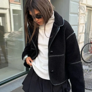 Svart teddyjacka med knappar - Intresse koll!!!! Snygg svart teddyjacka med knappar och kontrastsömmar. Jackan har en croppad passform och långa ärmar, perfekt för kyligare dagar. Den är både stilren och mysig, vilket gör den till ett måste i garderoben.