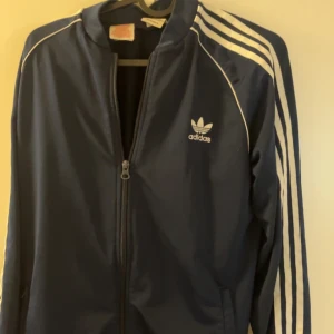 Svart träningsjacka från Adidas - Snygg svart träningsjacka från Adidas med klassiska vita ränder längs ärmarna och dragkedja framtill. Perfekt för sportiga aktiviteter eller som en stilren vardagsjacka.