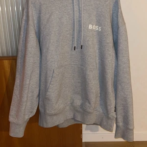 Grå hoodie från BOSS - Säljer en stilren grå hoodie från BOSS med justerbar dragsko i huvan och ribbade muddar. Perfekt för en avslappnad look. BOSS-loggan är tryckt på bröstet.