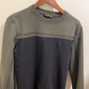 Grå och olivgrön tröja från Emporio Armani - Snygg tröja från Emporio Armani i en kombination av grått och olivgrönt. Tröjan har långa ärmar och ett diskret märke på bröstet. Den har också textdetaljer längs ärmarna och bröstet.