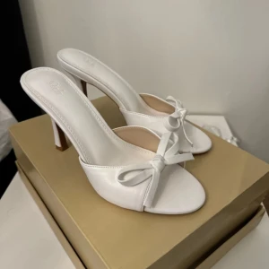 Vita klackskor med rosett - Eleganta vita klackskor med öppen tå och en söt rosett framtill. Perfekta för en stilren look. Helt nya aldrig använda.