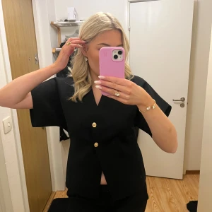 Svart kortärmad topp från Zara - Helt ny populär svart topp från Zara med korta ärmar och v-ringning. Toppen har dekorativa guldknappar framtill och en stilren design. Prislappen är fortfarande kvar.