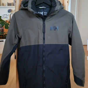 Skidjacka, Helly Hansen Traverse  - Säljer en riktigt fin tvåfärgad jacka från Helly Hansen i olivgrön och svart. Jackan har en praktisk huva och dragkedja framtill. Från 12 år, men funkar i flera åldrar pga sin justerbara längd, nedtill och i ärmslutet. Perfekt för skidbacken! Läs mer: https://www.skidresor.com/skidshop/skidjackor-barn/helly-hansen-traverse-skidjacka-junior-svart/
