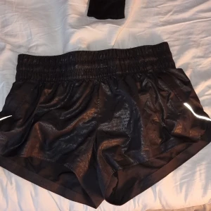 Korta svarta träningsshorts - Säljer ett par korta, svarta shorts från H&M med elastisk midja och reflekterande detaljer. Perfekta för träning eller en aktiv dag. Materialet är syntetiskt och aningen glansigt. 