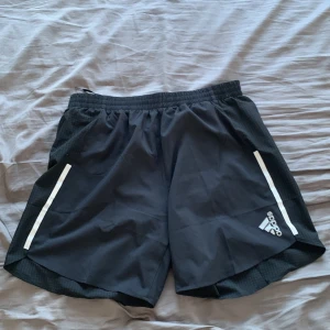 Svarta träningsshorts från Adidas - Säljer ett par svarta träningsshorts från Adidas, tillverkade av Parley Ocean Plastic. Shortsen har en elastisk midja och vita detaljer på sidorna. Perfekta för löpning med AEROREADY-teknologi för att hålla dig torr och bekväm.
