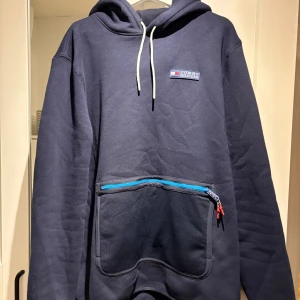 Mörkblå hoodie från Tommy Hilfiger - Säljer en snygg mörkblå hoodie från Tommy Hilfiger med en stor ficka framtill och dragkedja i blått. Hoodien har en klassisk design med snören vid huvan och en liten logga på bröstet.