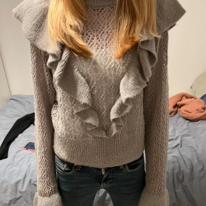 Grå stickad tröja med volangdetaljer - Säljer en snygg grå stickad tröja med volangdetaljer framtill och på ärmarna. Tröjan har ett fint mönster och långa ärmar, perfekt för kyligare dagar. Passar bra till jeans eller kjol för en stilren look.