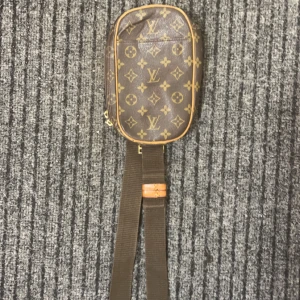 Brun axelväska från sLv - Säljer en snygg brun axelväska från SLv med det klassiska monogrammönstret. Väskan har en justerbar rem och dragkedja för säker förvaring. Perfekt för att bära dina nödvändigheter med stil.