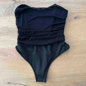 Svart body - Elegant svart body med rynkade detaljer som ger en smickrande passform. Den är även axelbandslös men håller upp sig bra. Bodyn är perfekt och stilren för alla händelser. Finns inga defekter och använd fåtal gånger men har varit mestadels i garderoben i 1 år🥰💓