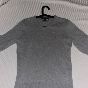 Grå tröja från Tommy Hilfiger - Säljer en stilren grå tröja från Tommy Hilfiger i  bomull. Tröjan har en v-ringad design och en  broderad logga på bröstet. Perfekt för en avslappnad och snygg look på en lite kyligare dag. Inga fläckar, hål eller liknande👍                           Tror nypris ligger på runt 1000 kr.                                  Mitt pris: 349. Kan tänka mig att gå ner lite i pris vid snabb affär.                                                        Bara att höra av sig vid frågor😃