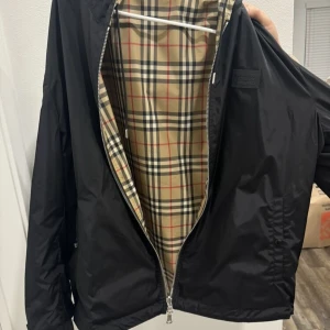 Burberry Reverseble Jacka - Snygg svart jacka från Burberry med klassiskt rutigt foder. Jackan har en dragkedja framtill och en huva för extra skydd. Perfekt för en stilren look. Skyddar från vind och regn