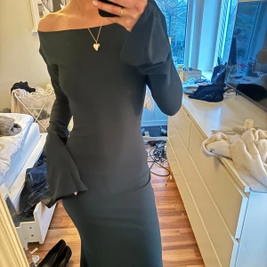 Grön långklänning med öppen rygg - Elegant mörkgrön långklänning med offshoulder-design och långa ärmar. Klänningen har en stilren öppen rygg med knytband i nacken, vilket ger en sofistikerad look. Nypris 945kr