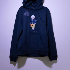 Polo Ralph Lauren Hoodie - Helt ny Polo Ralph Lauren Hoodie.Tröjan har en nallebjörn på framsidan och en klassisk känguruficka och justerbar huva. Perfekt för en avslappnad stil.