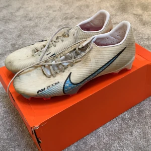 Nike Mercurial fotbollsskor - Säljer ett par vita Nike Mercurial fotbollsskor med blå och rosa detaljer. Skorna har snörning och en modern design med Air Zoom-teknologi för optimal prestanda på planen.dom ser smutsiga ut men man kan tvätt dom jätte lätt. Priset kan diskuteras 