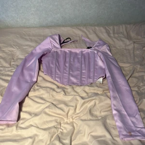 Lila korsett från Missguided - Snygg lila korsett från Missguided med långa ärmar och dragkedja bak. Perfekt för en trendig look. Tillverkad i ett glansigt material som ger en lyxig känsla. Köpt och använd i USA en gång, aldrig använt sedan dess. Har dock en fläck på ärmen (se bild) 