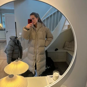Beige lång dunjacka - Säljer en beige lång oversize dunjacka med huva och dragkedja. Perfekt för kalla dagar med sin pufferdesign som håller dig varm och bekväm. 