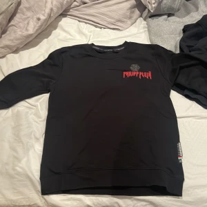 Svart tröja från Philipp Plein - Säljer en svart tröja från Philipp Plein med röd logotyp på bröstet och en liten etikett på sidan. Tröjan har rund hals och är långärmad, perfekt för en stilren look.