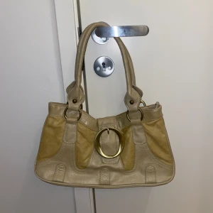 Beige handväska med gulddetaljer - Snygg beige handväska med stora guldringar som detaljer. Väskan har två rejäla handtag. Insidan är fodrad med ett mönstrat tyg och har en praktisk innerficka med dragkedja. Perfekt för att bära dina dagliga nödvändigheter med stil.