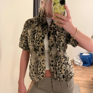 Leopardmönstrad kortärmad pälsjacka - Snygg och trendig leopardmönstrad pälsjacka med korta ärmar. Perfekt för att ge din outfit en extra edge. Går att stänga igen med ett spänne som sitter fast på jackan.