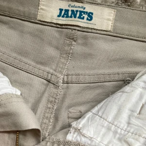 Beige jeans från Calamity Jane's - Snygga beige jeans från Calamity Jane's med klassisk femficksdesign. Byxorna har en rak passform och är perfekta för grounge stil. Tillverkade i ett bekvämt material som passar till många olika tillfällen.