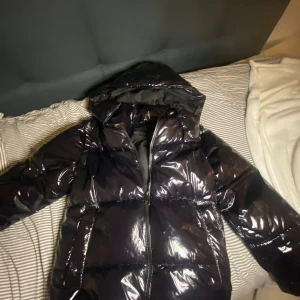 Svart glansig pufferjacka från Zara - Snygg svart pufferjacka från Zara med glansig finish. Jackan har en dragkedja framtill och en mysig huva. 
