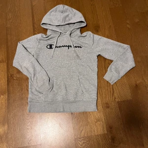 Grå hoodie från Champion - Säljer en klassisk grå hoodie från Champion med deras ikoniska logga tryckt på bröstet. Tröjan har en bekväm passform med långa ärmar och en praktisk huva med dragsko. I mycket bra skick och bär storleken S.