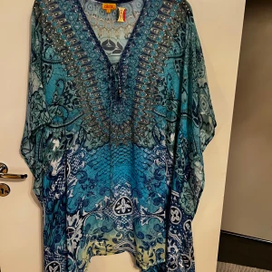 Blå mönstrad kaftan från Akaru - Säljer en vacker blå kaftan från Akaru med ett detaljerat mönster i olika nyanser av blått och grönt. Kaftanen har en v-ringad hals med knytband och löst sittande passform. Perfekt för en avslappnad och stilfull look.