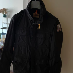 Svart jacka från Parajumpers - Snygg svart jacka från Parajumpers med emblem på ärmen. Är en typ av fieldjacka. Jackan har flera fickor och en hög krage med en gul detalj vid halsen. Perfekt för blåsigare dagar. Nypris 6000kr. Inga hål eller liknande.