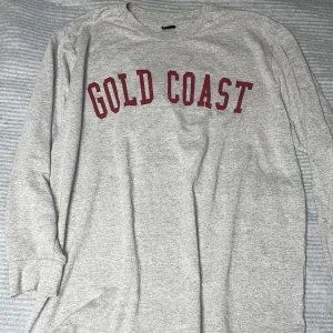Grå tröja från Gina Tricot med text - Säljer en grå långärmad tröja från Gina Tricot med texten 'GOLD COAST' i rött på framsidan. Perfekt för en avslappnad stil.