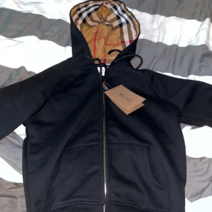 Svart huvtröja från Burberry - Säljer en stilren svart huvtröja från Burberry med dragkedja och klassiskt rutigt mönster i huvan. Perfekt för en avslappnad och trendig look. Storlek L men är liten i storleken.