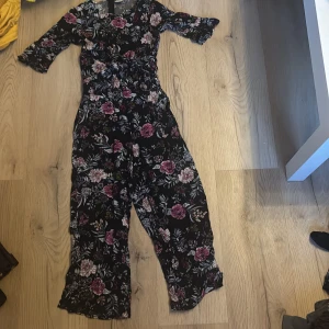 Blommig jumpsuit från Lindex - Snygg blommig jumpsuit från Lindex i svart med rosa och vita blommor. Den har trekvartsärmar och en knytning i midjan för en fin passform. Perfekt för en stilren look!
