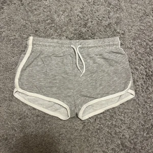 Grå bomullsshorts med snörning - Säljer ett par bekväma grå shorts i bomull med vit snörning i midjan. Perfekta för en avslappnad dag hemma. De har en elastisk midja och vita detaljer längs kanterna.