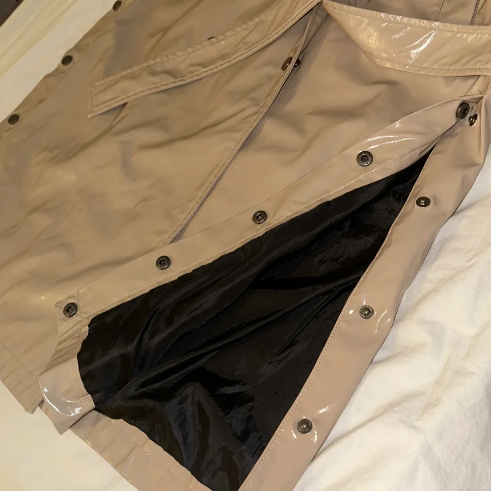 Snygg beige trenchcoat från Veromoda i glansigt vattentätt material. Jackan har långa ärmar och knappar framtill samt ett bälte i midjan för en stilren look. Perfekt för att ge din outfit en trendig touch.. Takit.