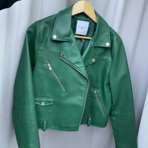 Grön skinnjacka från MNG - Snygg grön skinnjacka från MNG med asymmetrisk dragkedja och stilrena detaljer. Jackan har långa ärmar och flera praktiska fickor med dragkedja och knappar. Perfekt för en cool och trendig look.