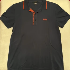 Mörkblå/marinblå pikétröja från Hugo Boss - Använd 1 gång. Snygg mörkblå pikétröja från Hugo Boss i slim fit. Tillverkad av mjuk Pima bomull med orange detaljer på kragen och knäppningen. Perfekt för en stilren look.