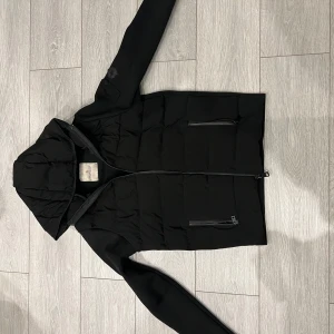 Svart cardigan från Moncler - Snygg svart cardigan från Moncler med huva och dragkedja. Jackan har en stilren design med Moncler-logga på ärmen och praktiska fickor med dragkedja. Perfekt för kyligare dagar.