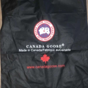 Svart dunjacka från Canada Goose - Säljer en svart dunjacka från Canada Goose med huva och broderad logga på ärmen. Jackan har en stilren design med dragkedja och knappar framtill. Perfekt för kalla dagar.